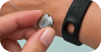 A woman holds a pebble away from the FIVE.FIT band, with her skin visible through the empty opening.